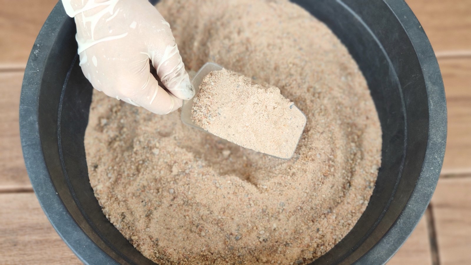 A gloved hand holds a small shovel filled with fine, light beige sand above a bucket brimming with smooth, evenly textured sand.
